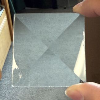 Close-up of a small square metal panel with a reflective protective film creating an X pattern, held between two fingers.